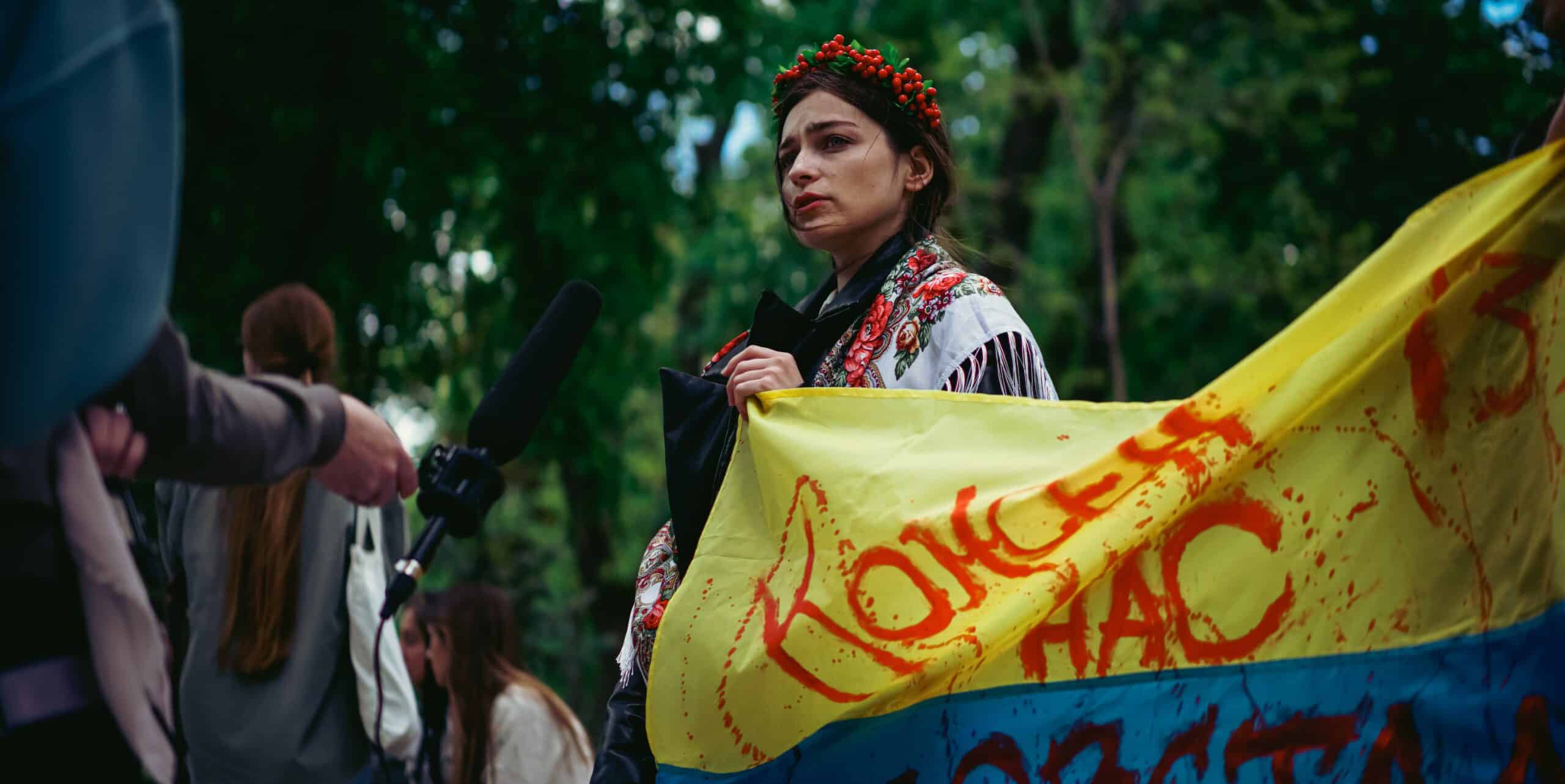 A woman holds a bloodstained flag of Ukraine in her hands, dressed in traditional Ukrainian clothing, protesting the war. Someone is holding a microphone close to her.